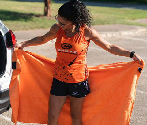Runner in an Orange Mud singlet and shorts holding open an orange Transition Wrap Extreme changing towel beside her car's open hatch in a parking lot