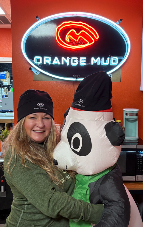Person hugging a mascot in front of an Orange Mud neon sign.